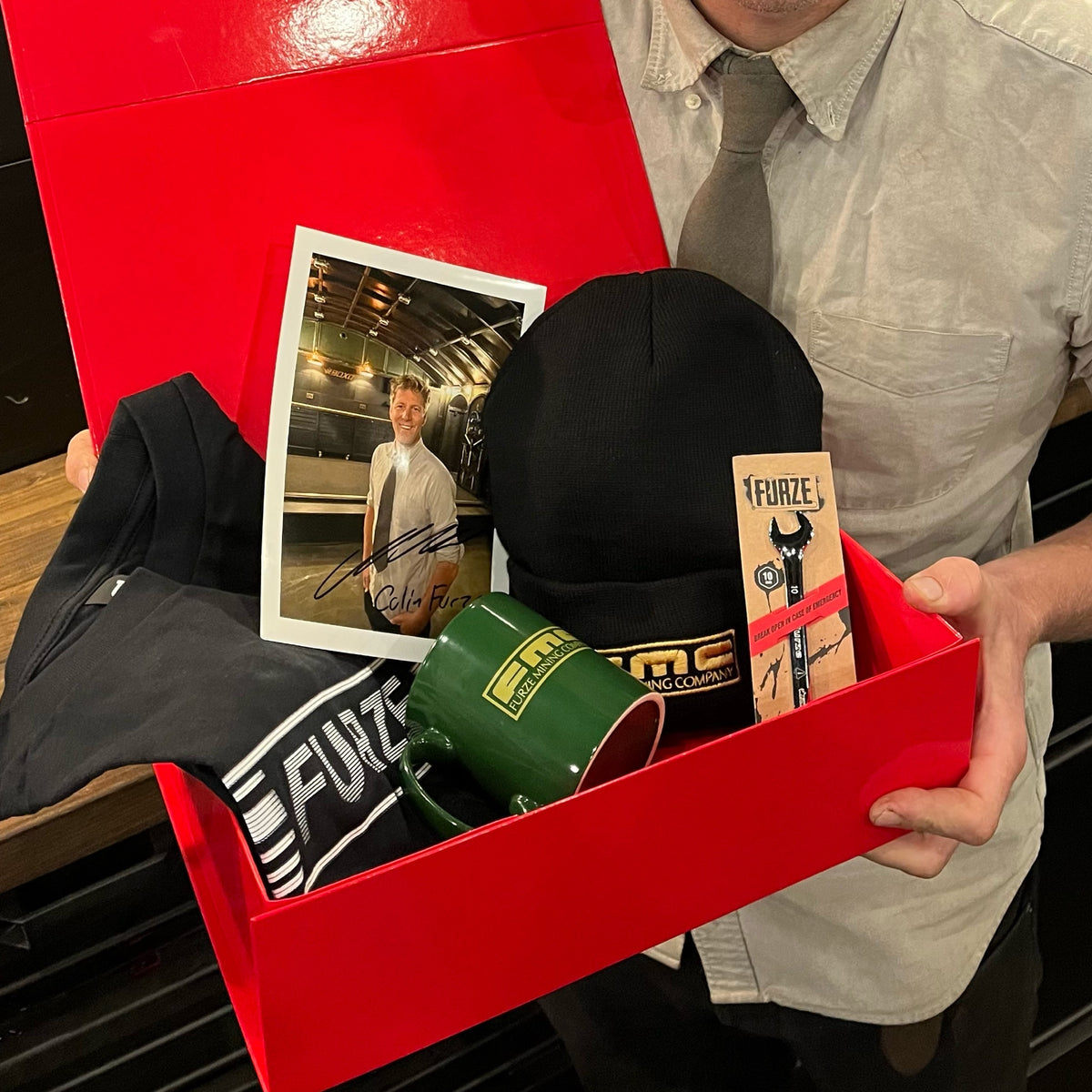 Colin Furze holding the open FMC Bundle box with signed photo, beanie, mug, T-shirt and spanner