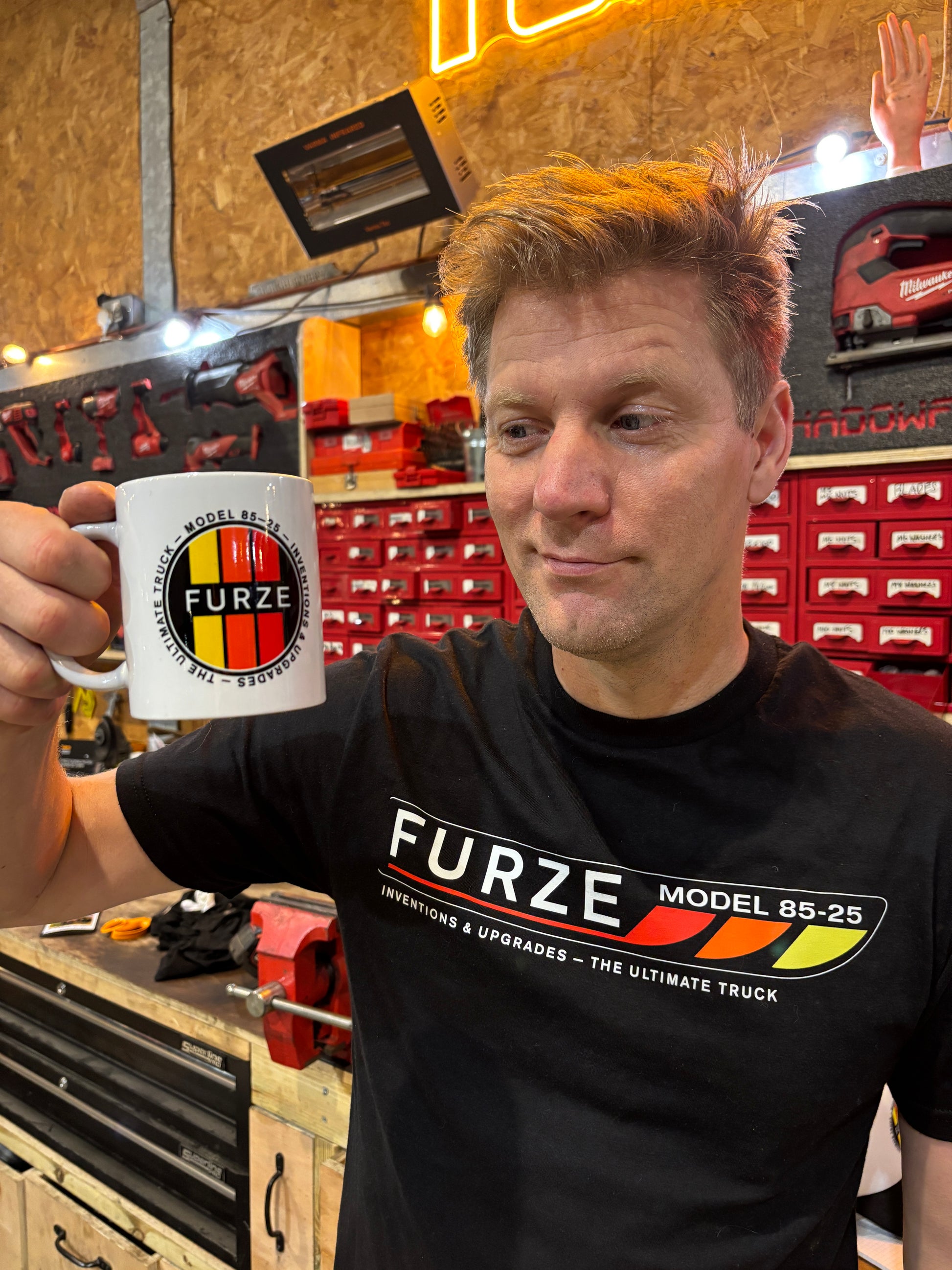 Colin Furze wearing a black 'Furze Model' t-shirt holding a white mug with the same brand logo in a workshop setting.