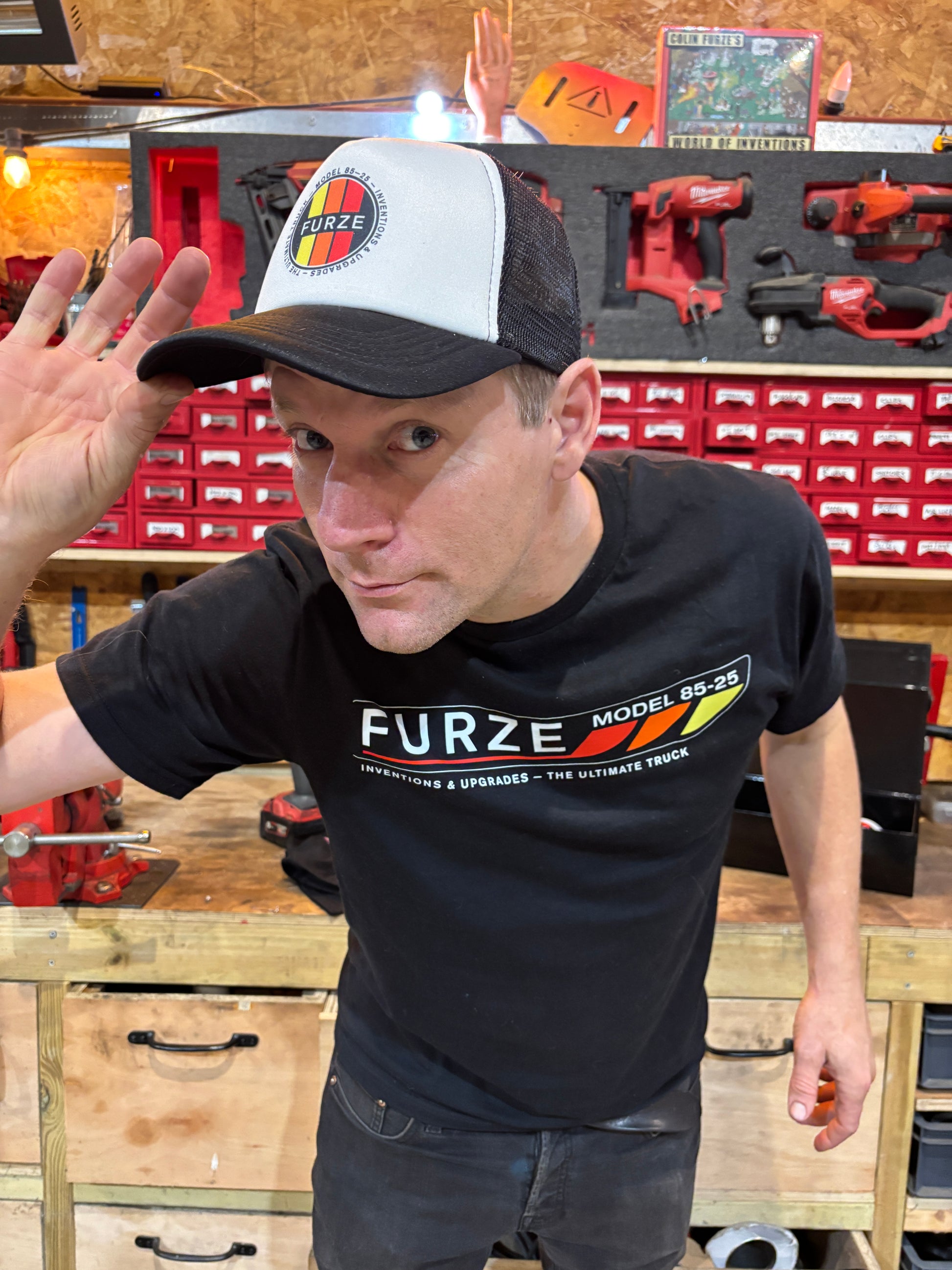 Colin Furze wearing a black 'Furze' t-shirt in a workshop setting with tools and equipment.