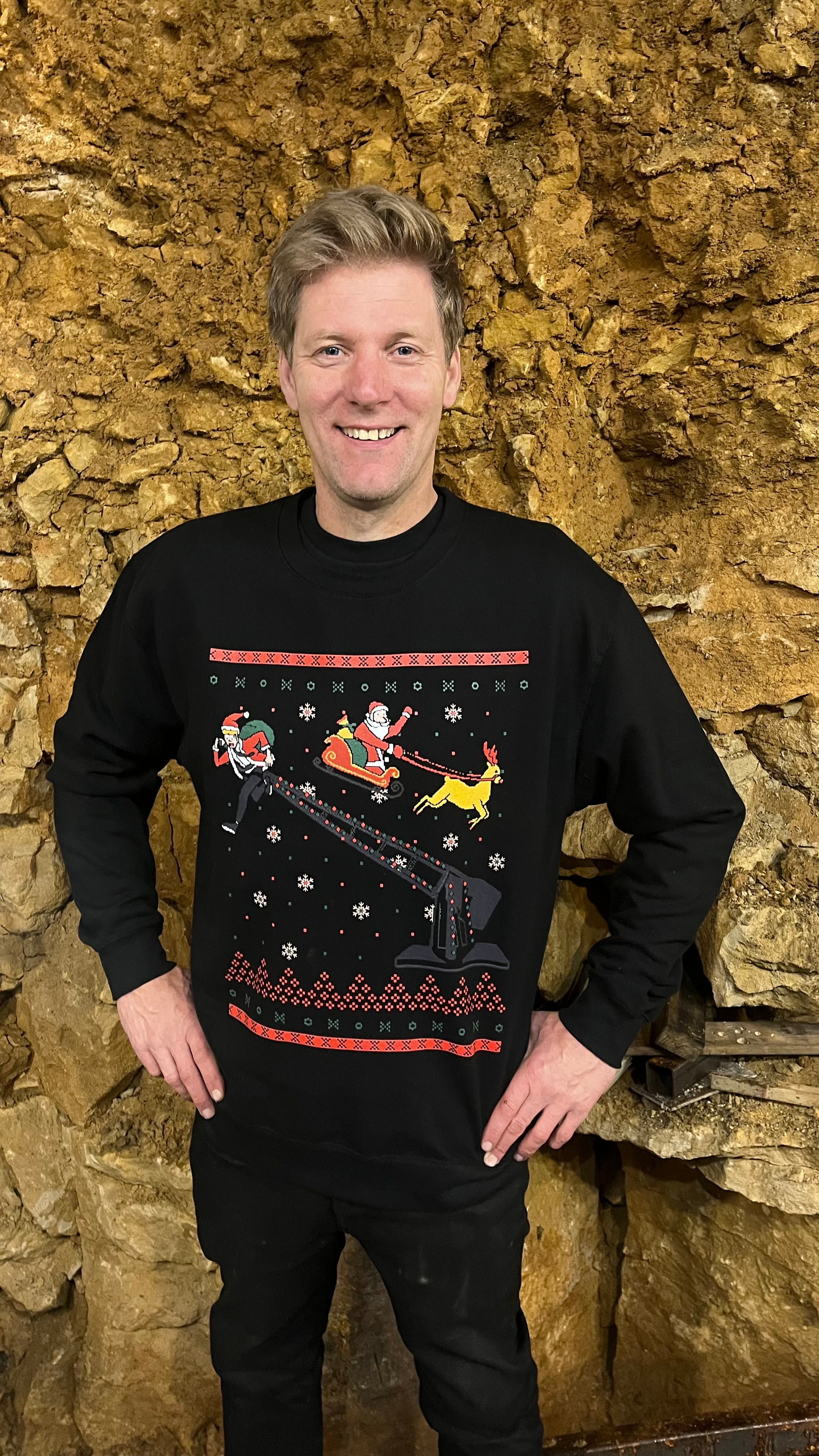 Colin standing in front of a rocky background, wearing a black Christmas-themed sweater with Santa Claus and a reindeer on the front.