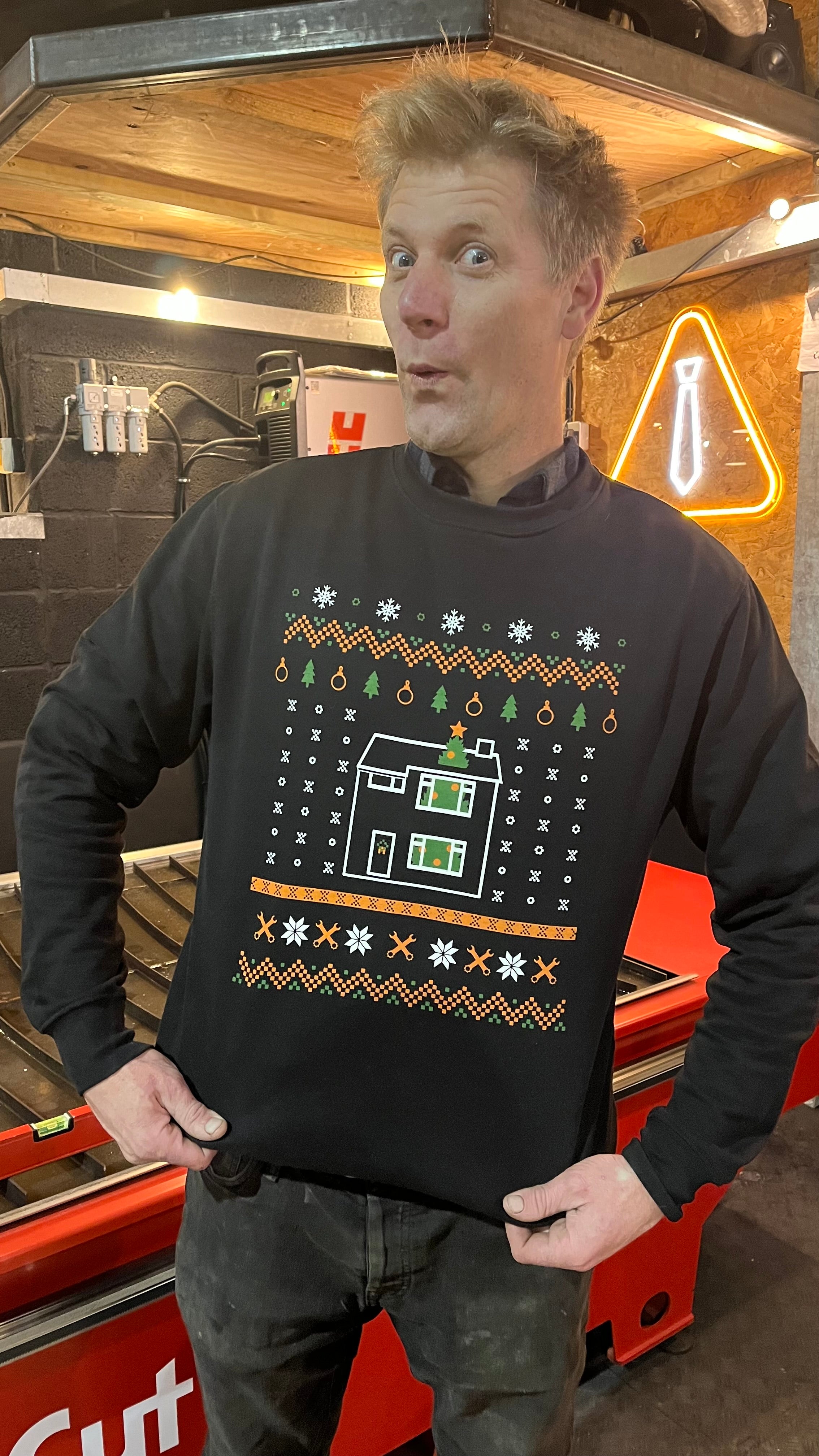 Colin standing and posing with a Christmas-themed jumper featuring a large tree and house design.
